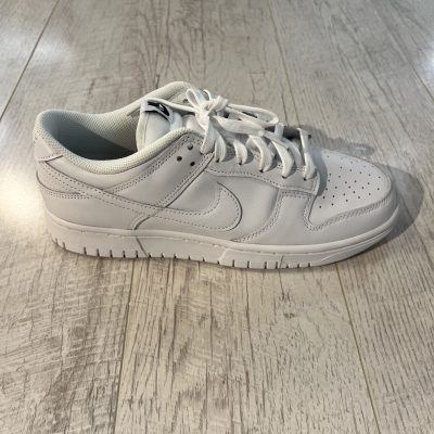 Vu de coté de la Nike dunk low triple white. Une couleur blanche sur l'ensemble de la paire. La matière est dur cuir. Des lacets blancs.