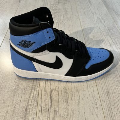 Vu de coté de la Nike Air Jordan 1 High OG University Blue UNC Toe. Du bleu et du noir ainsi que du blanc. Un swoosh nike noir. Des lacets noirs.