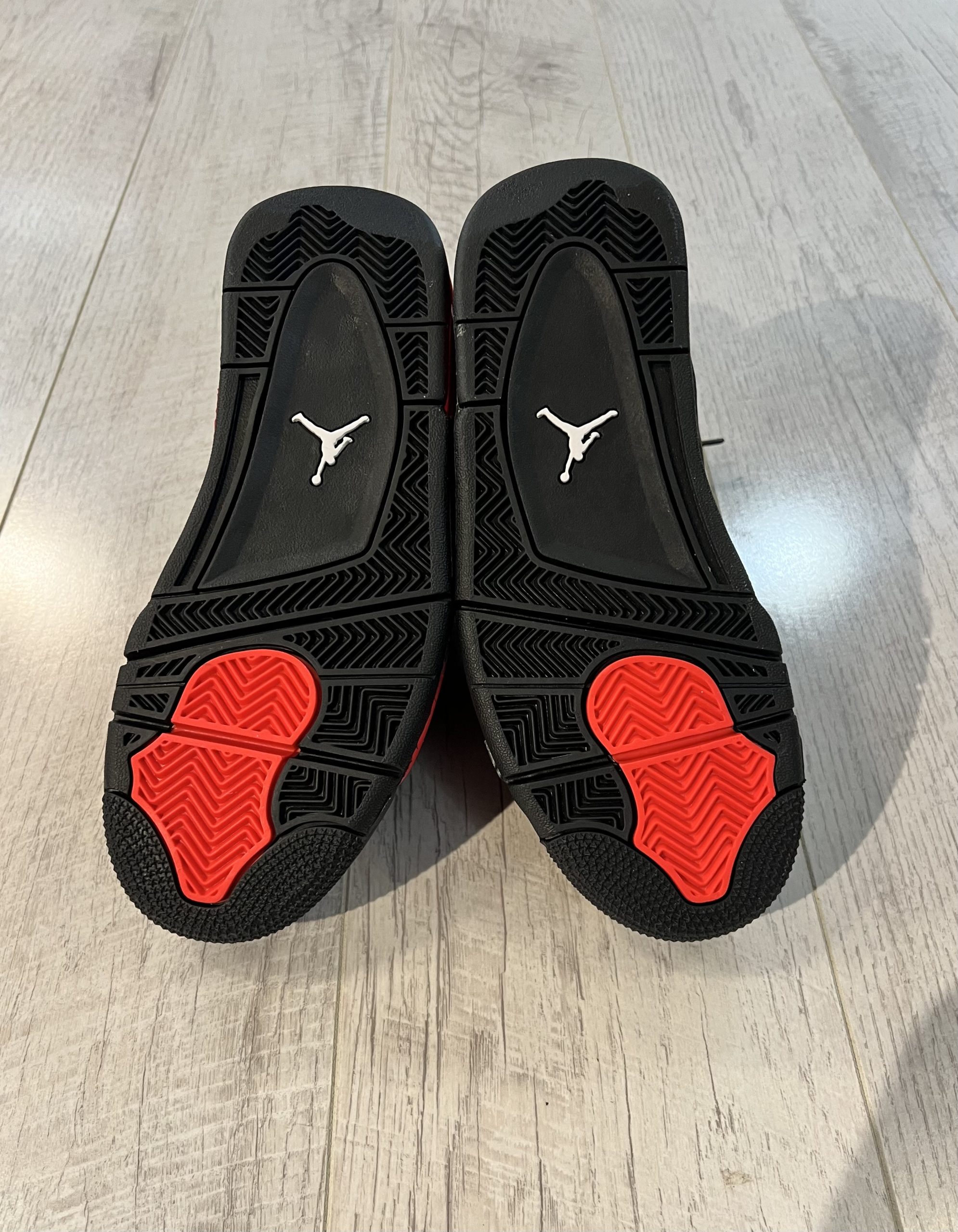 nike-air-jordan-4-red-thunder.jpeg Vu du dessous de la Nike Air Jordan 4 Red Thunder. Du. noir domine la paire, une touche de rouge sur l'avant de la semelle. Un logo jordan blanc au milieu.