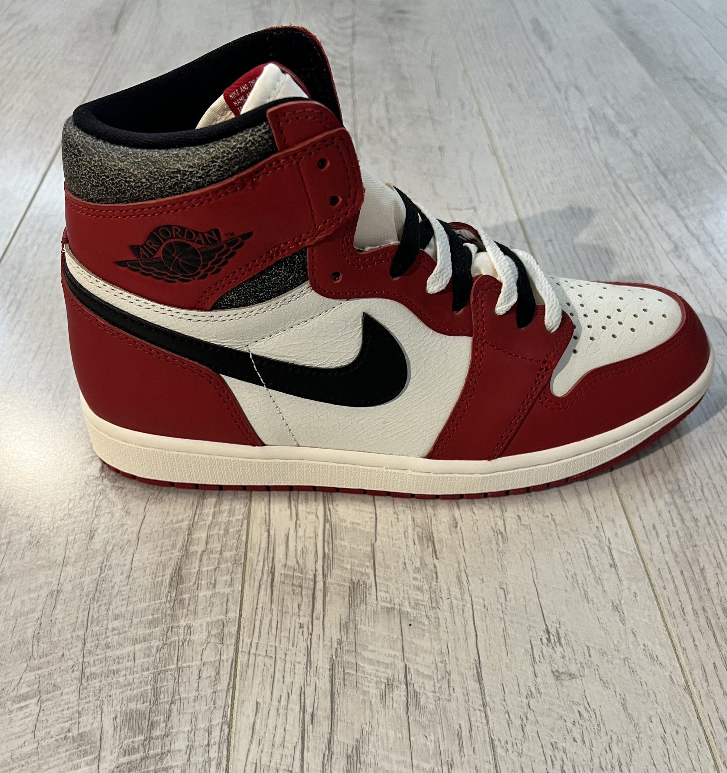nike-air-jordan-1-high-og-lost-and-found.jpeg Vu de coté de la Nike Air Jordan 1 High OG Lost and Found. Le rouge et le blanc dominent. Un swoosh nike noir, une languette beige, des lacets noirs et blancs. L'intérieur de la paire est noir.