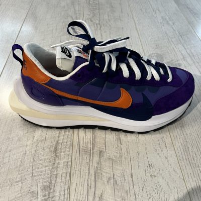 Vu de coté de la Nike Sacai VaporWaffle Dark Iris. Un violet foncé et clair dominent en daim. Un signe nike orange et noir. Une semelle plus epaisse blanche et creme au talon.
