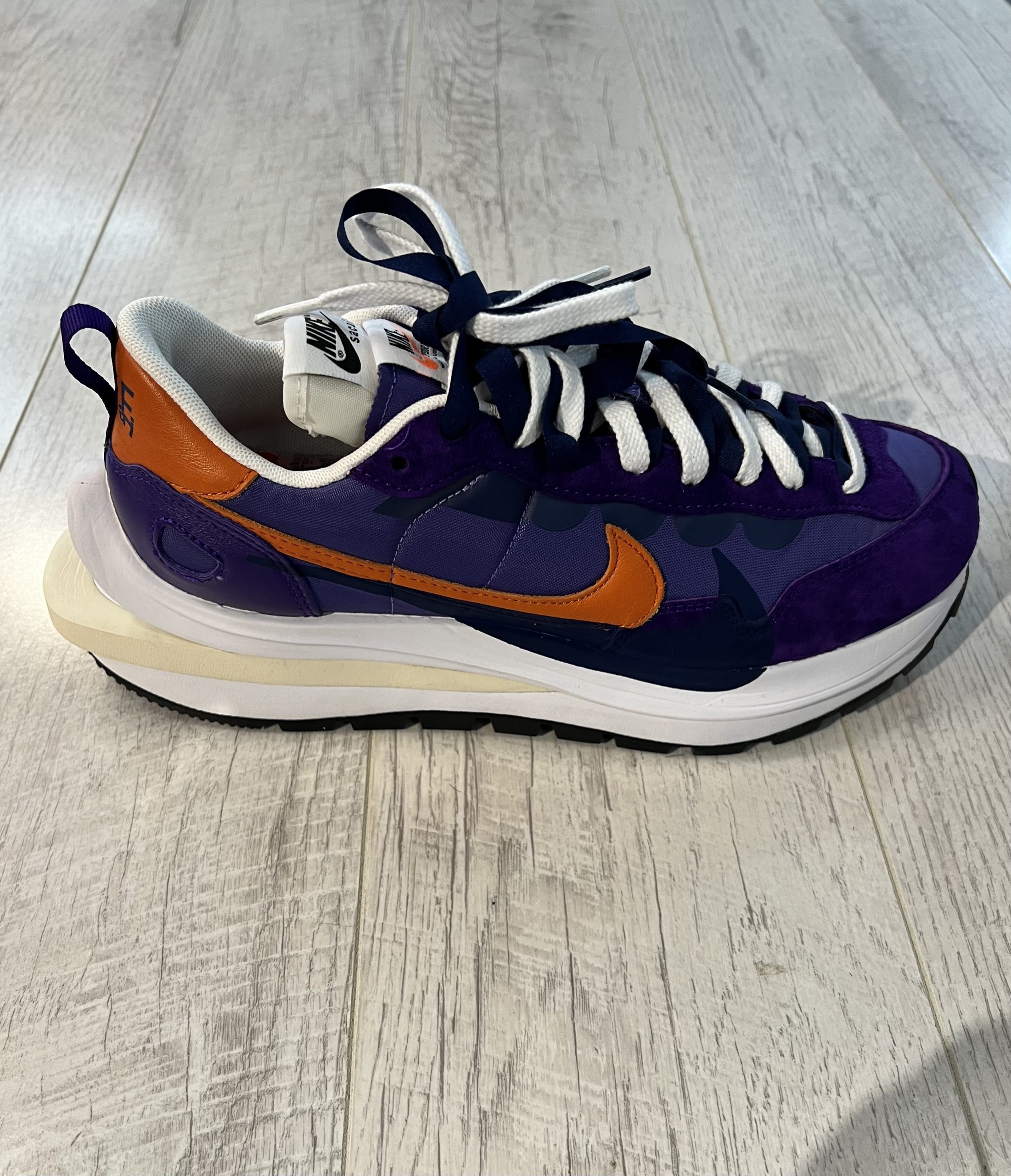 nike-sacai-vaporwaffle-dark-iris Vu de coté de la Nike Sacai VaporWaffle Dark Iris. Un violet foncé et clair dominent en daim. Un signe nike orange et noir. Une semelle plus epaisse blanche et creme au talon.