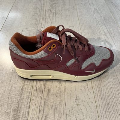 Vu de de coté de la paire de Nike Air Max 1 Patta Rush Maroon. Une couleur bordeaux et le signe Nike de la meme couleur. Du gris complète la paire. Une semelle beige et marron.