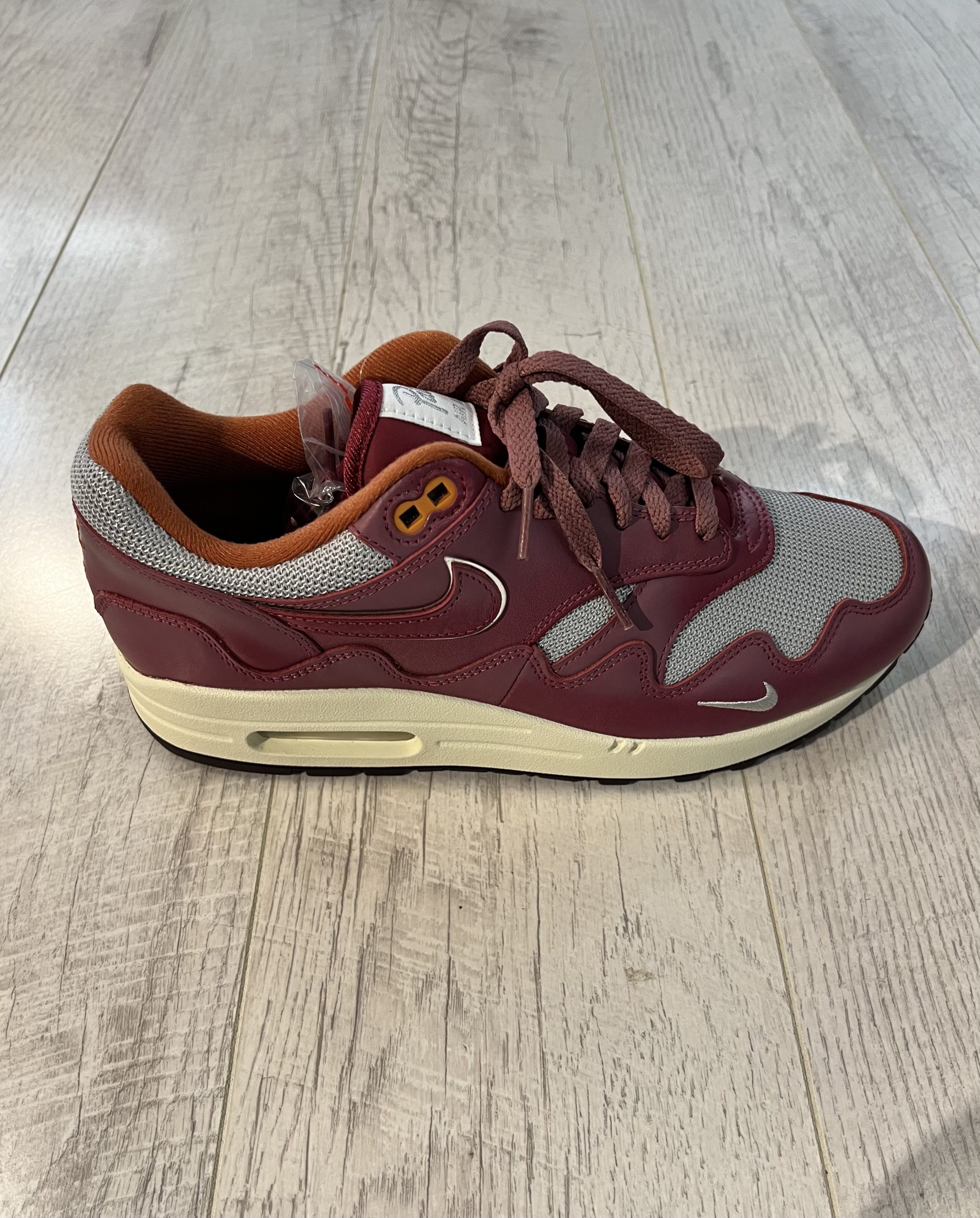 nike-air-max-1-patta-rush-maroom Vu de de coté de la paire de Nike Air Max 1 Patta Rush Maroon. Une couleur bordeaux et le signe Nike de la meme couleur. Du gris complète la paire. Une semelle beige et marron.