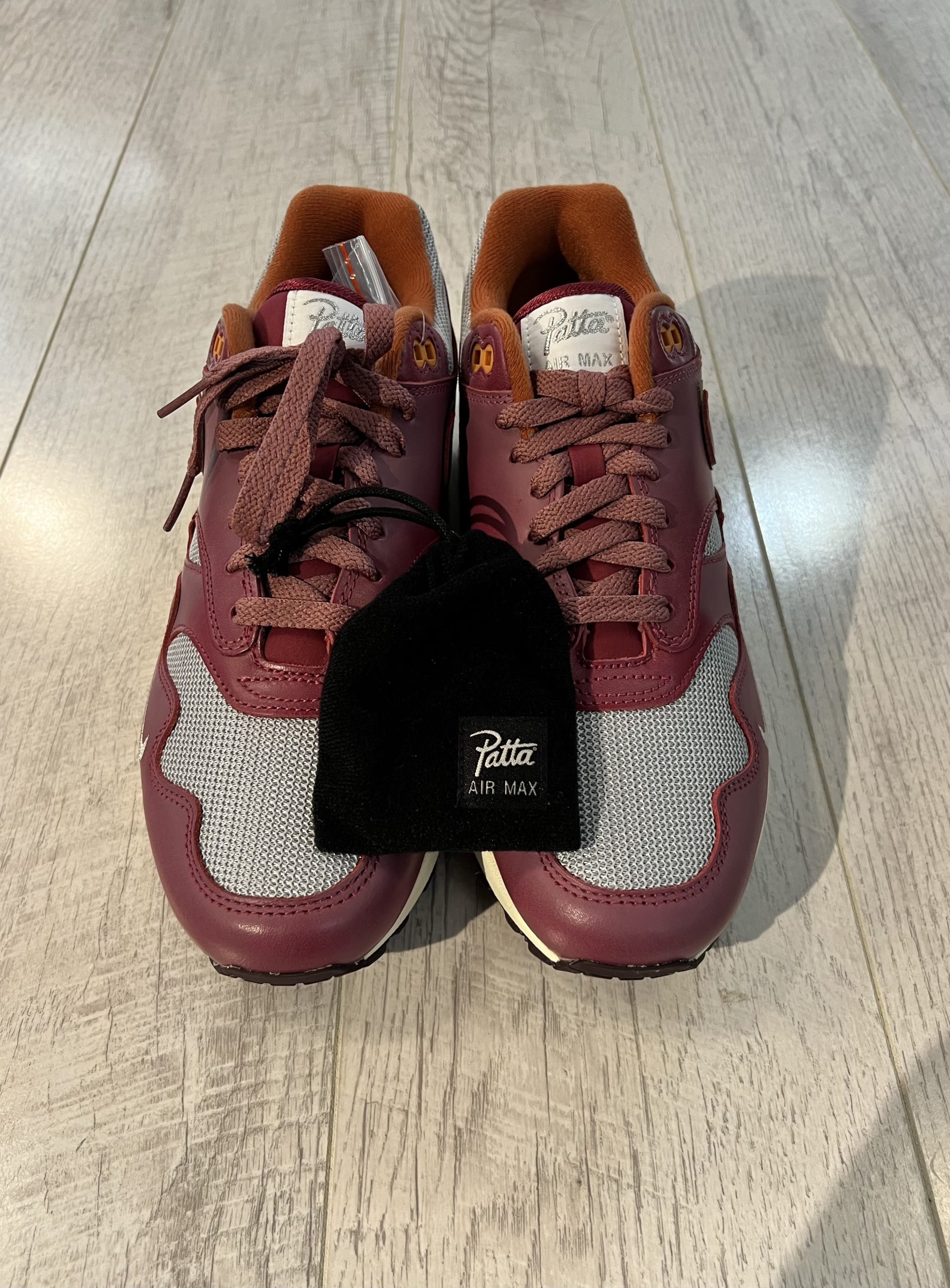 nike-air-max-1-patta-rush-maroom Vu du dessus de la Nike Air Max 1 Patta Rush Maroon. Du marron foncé en cuir et du gris en tissus dominent. Des lacets marrons foncés. Sur la languette le logo Patta en gris argenté sur un fond blanc. Une pochette noire avec des lacets à l'interieur.