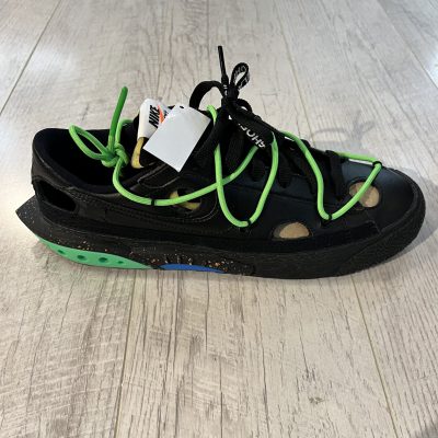 Vu de coté de la paire de Nike Blazer Low OFF White Black. Le noir domine la paire. Un lacet vert est sur le dessus de la paire. Des lacets noirs.