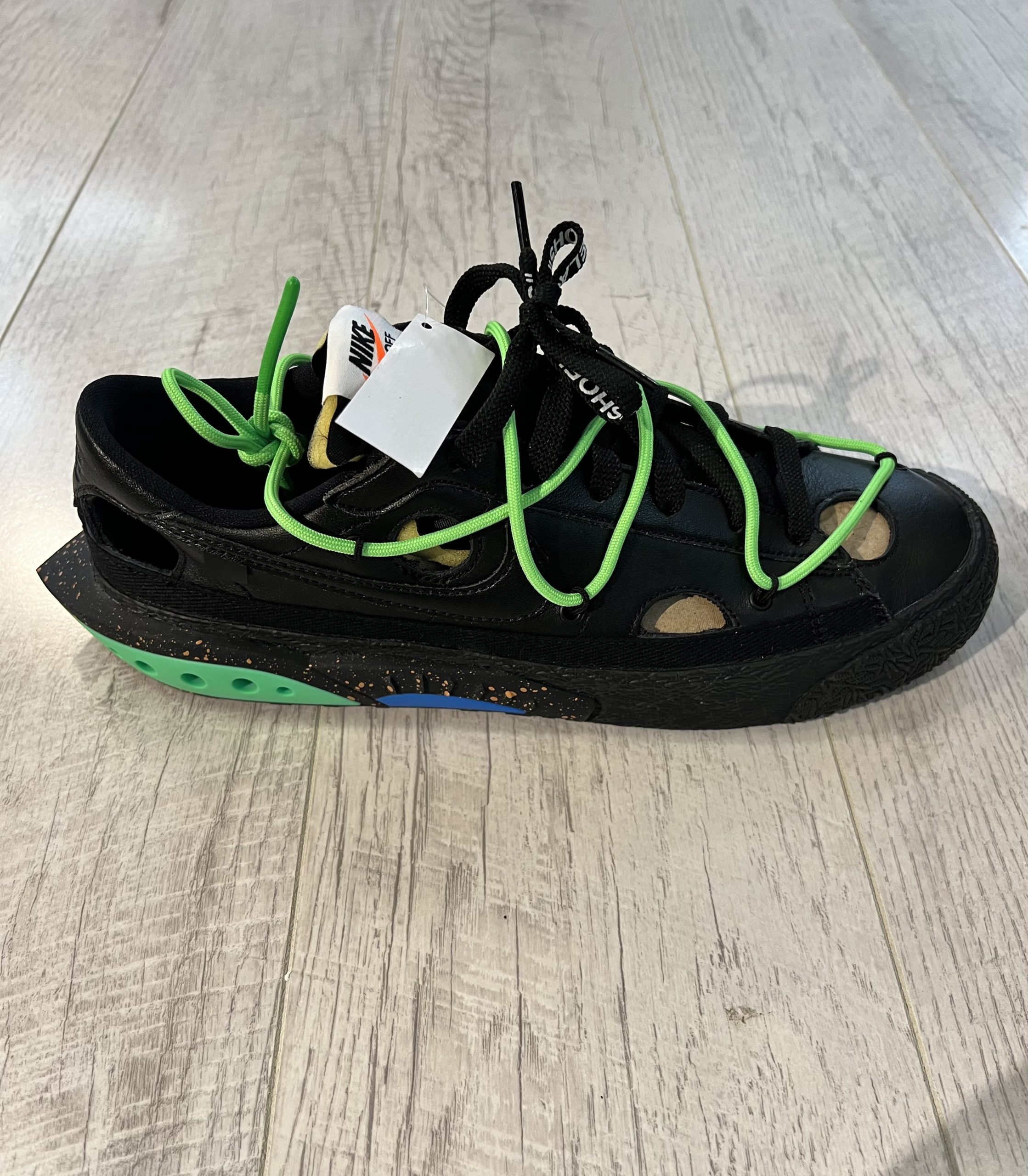 nike-blazer-low-off-white-black-.jpeg Vu de coté de la paire de Nike Blazer Low OFF White Black. Le noir domine la paire. Un lacet vert est sur le dessus de la paire. Des lacets noirs.