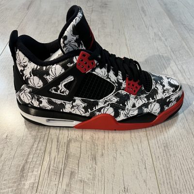 Vu de coté de la Nike Air Jordan 4 2018 Tattoo. Noir et blanc avec dessin sur du cuir. Oeillets rouge, semelle blanche rouge. Lacets noirs.